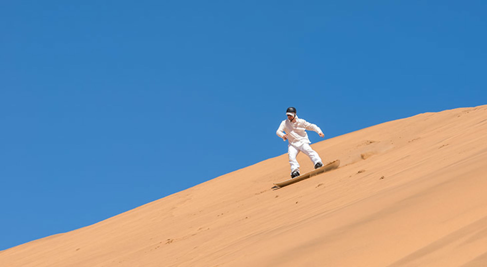 Man sandboardt van een hoge zandduin onder een helderblauwe lucht.