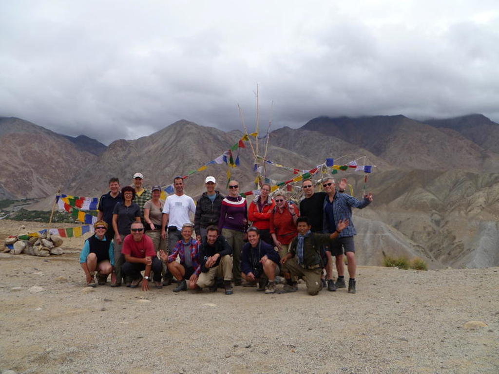 Groep wandelaars in Ladakh poseert bij gebedsvlaggen met berglandschap op de achtergrond.