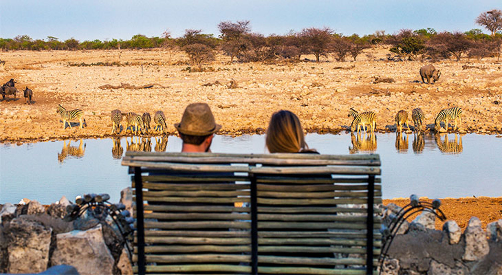 Twee reizigers kijken vanaf een bankje naar zebra’s bij een waterpoel op de savanne.
