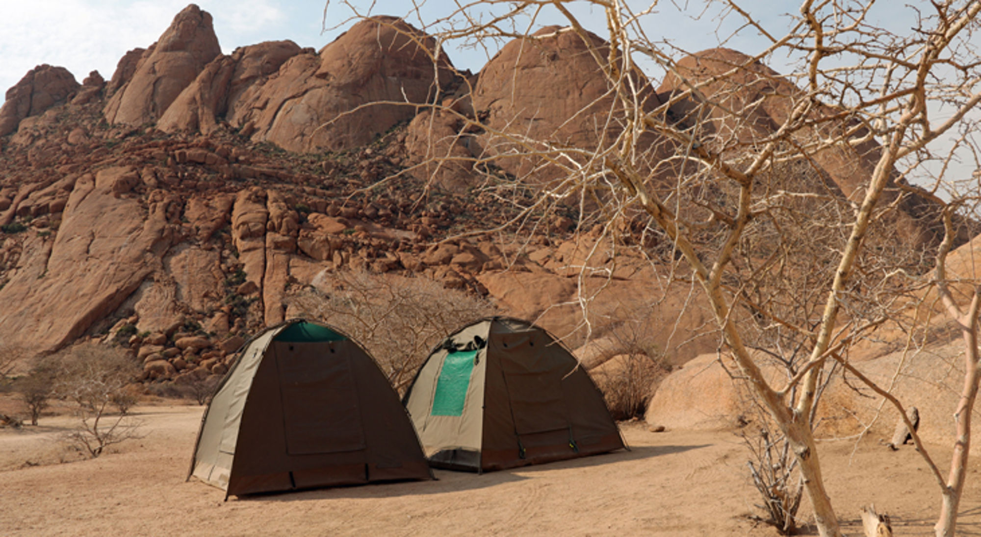 Kamperen bij Spitzkoppe in namibie Twee tenten op een zandvlakte bij rotsachtige bergen en kale struiken.
