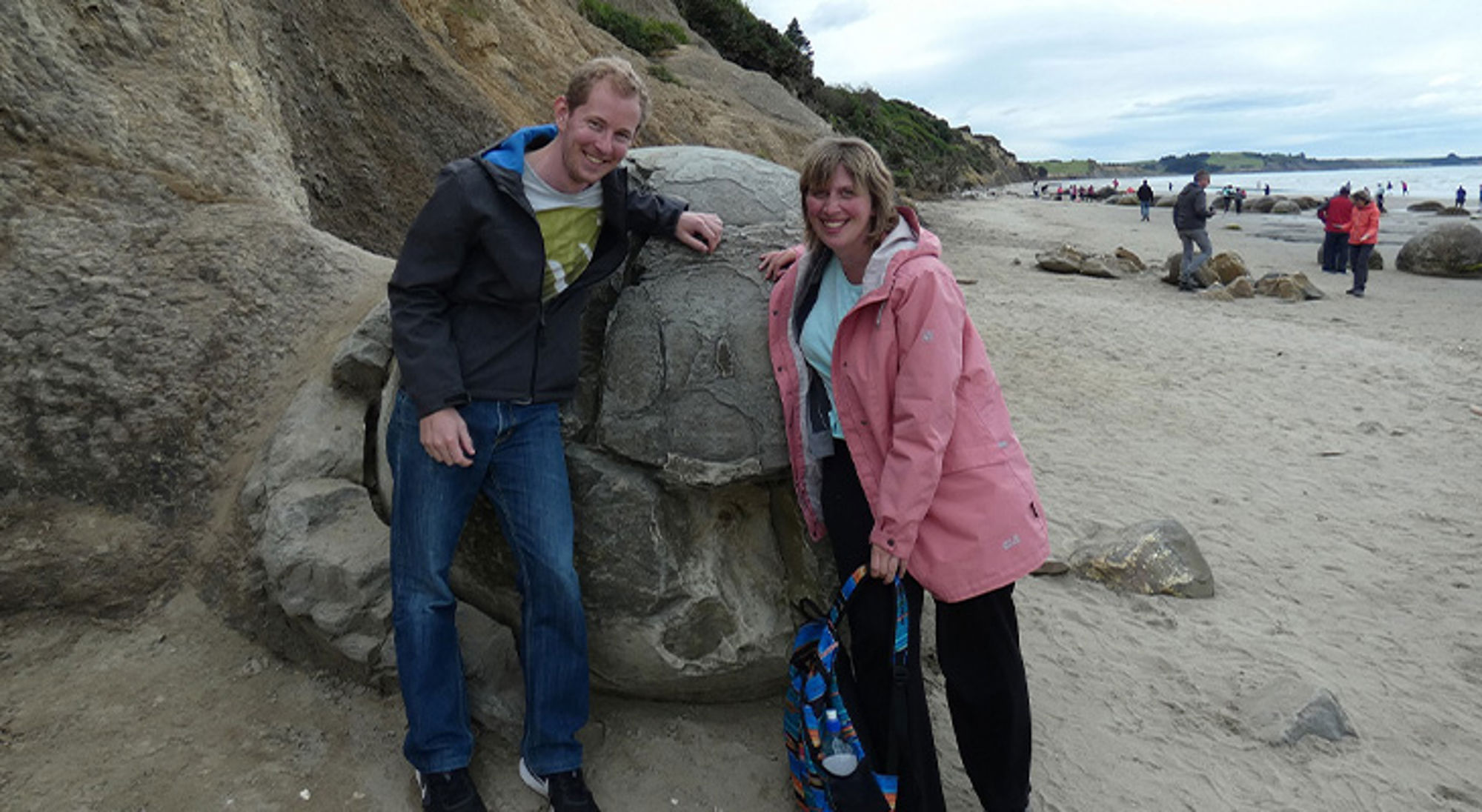 Twee reizigers poseren bij een grote rots op het strand, met andere bezoekers op de achtergrond.