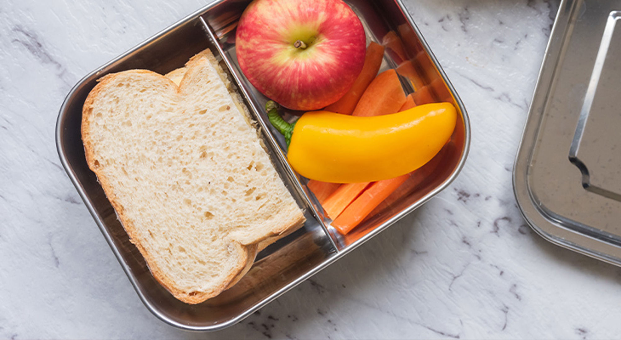 Broodtrommel vliegtuig RVS lunchbox met boterham, appel, wortel en gele paprika op een marmeren ondergrond.