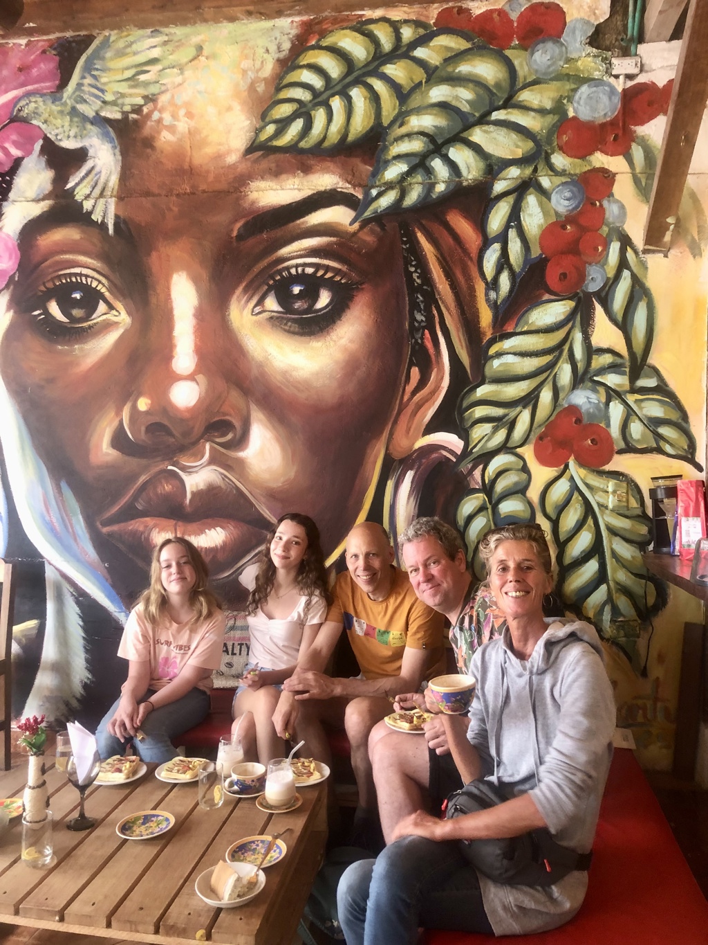 Familie geniet van koffie en taart in Colombiaans café met kleurrijke muurschildering op de achtergrond.