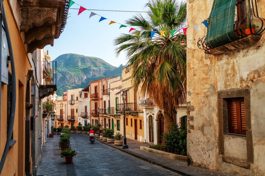 Straat in een dorp in West-Sicilië met kleurrijke vlaggetjes, palmbomen en bergen op de achtergrond.