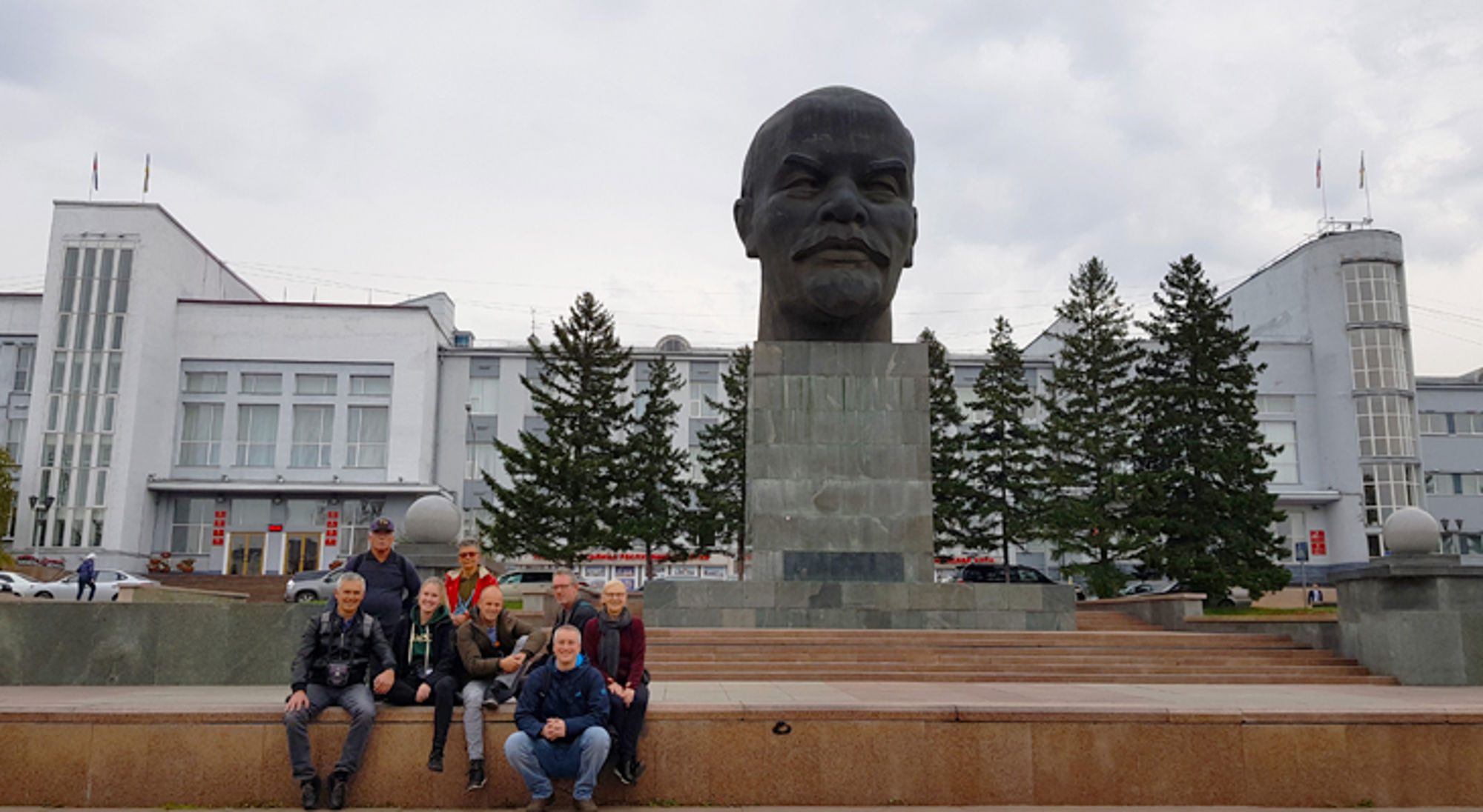 Reizigersgroep bij een groot Lenin-standbeeld op een stadsplein, met een wit overheidsgebouw op de achtergrond.