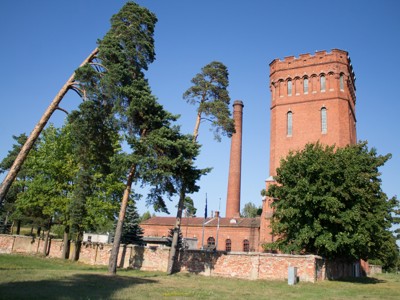 Rode bakstenen watertoren met schoorsteen, omringd door bomen langs de route Polen en Baltische Staten.