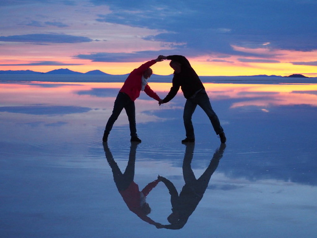 Twee reizigers vormen een hart met hun reflectie op de Salar de Uyuni zoutvlakte bij zonsondergang in Bolivia.