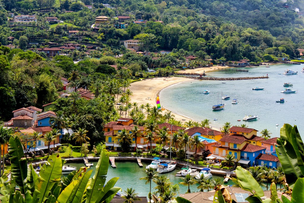 Uitzicht op een tropische baai in Brazilië met palmen, strand, kleurrijke huizen en boten op het water.