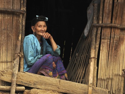 Lachende vrouw in kleurrijke kleding zit in een deuropening van een houten huis langs de route Laos & Cambodja.