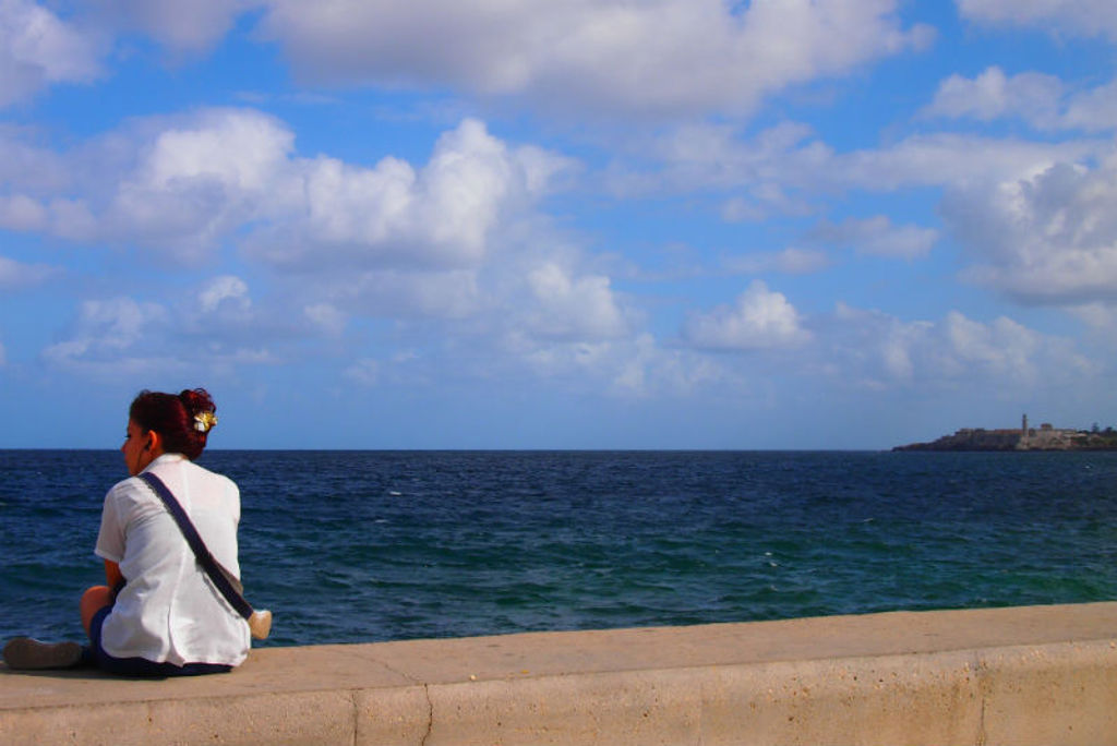 Vrouw zit op de Malecón in Havana en kijkt uit over zee onder een blauwe lucht met wolken.