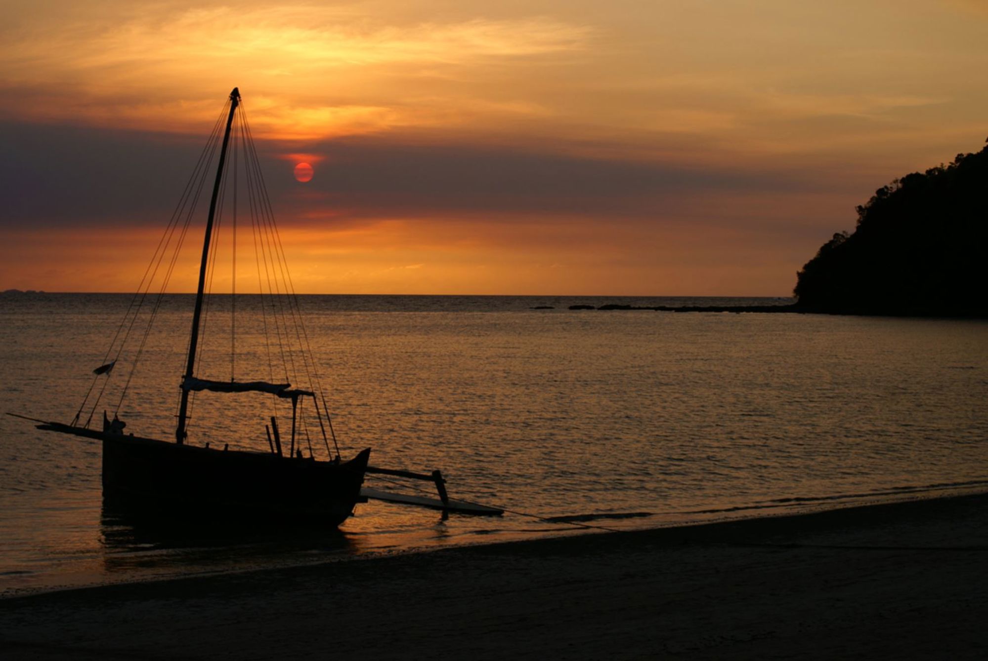 Zonsondergang aan de kust van Madagaskar met een traditionele zeilboot in silhouet op het strand.