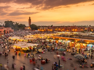 Djemaa el Fna-plein in Marrakech bij zonsondergang, met verlichte marktkramen en de Koutoubia-minaret op de achtergrond.