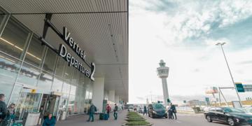 Vertrekhal Schiphol met bord “Departures 2” en reizigers met koffers bij de ingang.