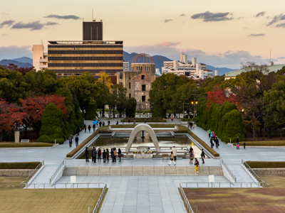 Atoombomkoepel in Hiroshima Peace Memorial Park met herdenkingsmonument en bezoekers bij zonsondergang.