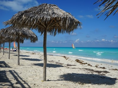 Rieten parasols op wit zandstrand met turquoise zee en zeilboot, Cuba.