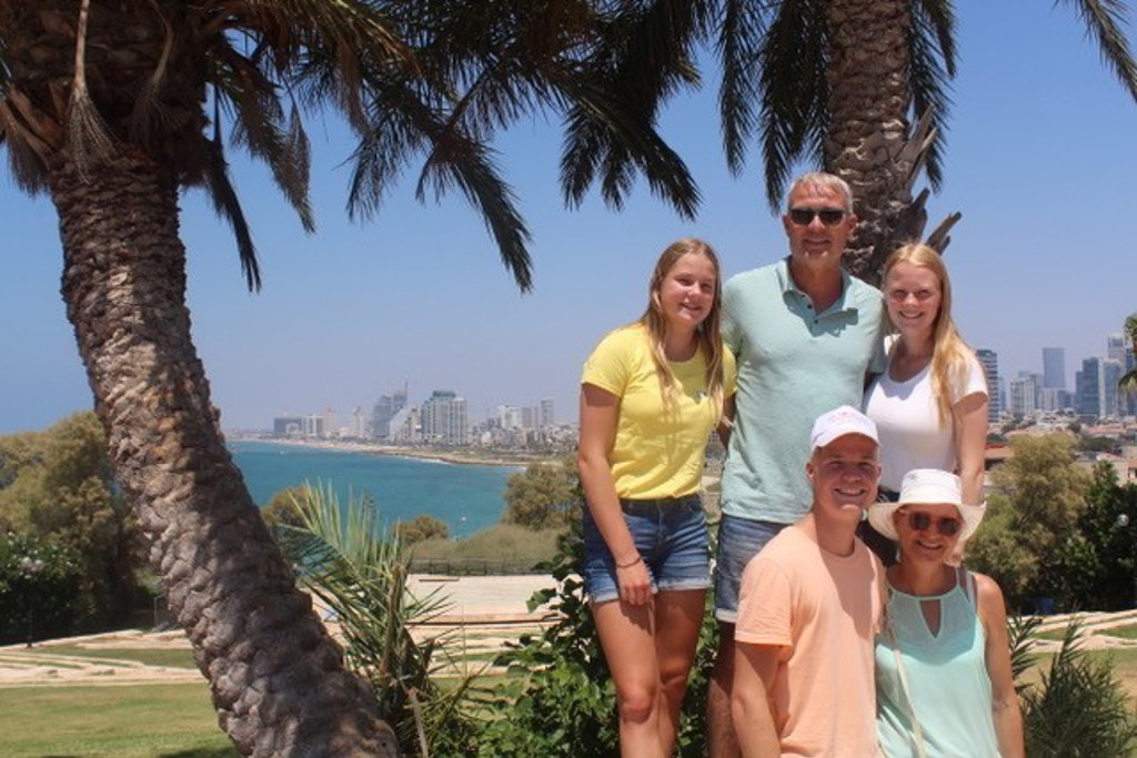 Gezin op familiereis in Israël aan de kust, met palmbomen en skyline op de achtergrond.
