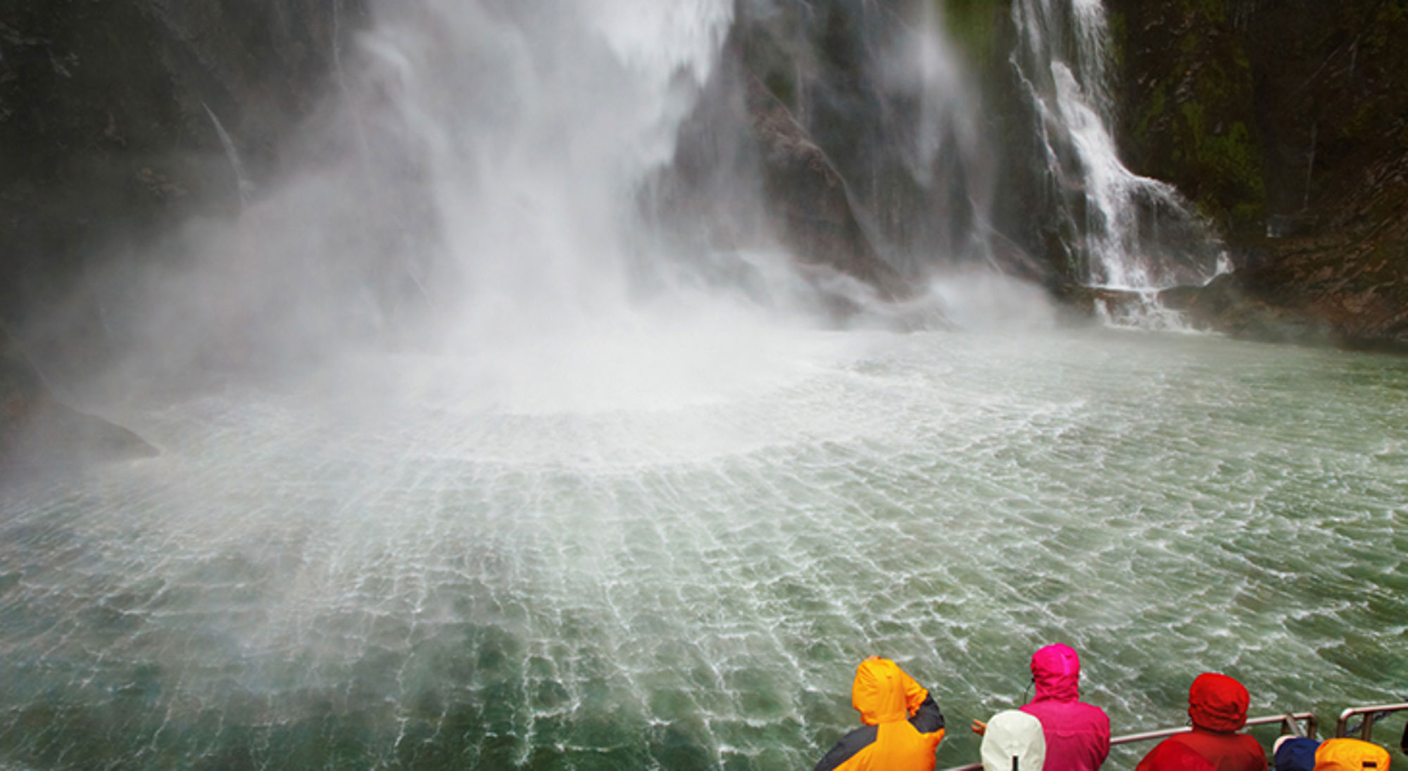 Milford Sounds in Nieuw-Zeeland Toeristen in regenjassen kijken vanaf een boot naar een krachtige waterval met opspattende nevel.