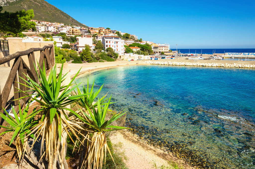 Kustplaats op Sardinië met helderblauwe baai, strand en haven, gezien vanaf een wandelpad.