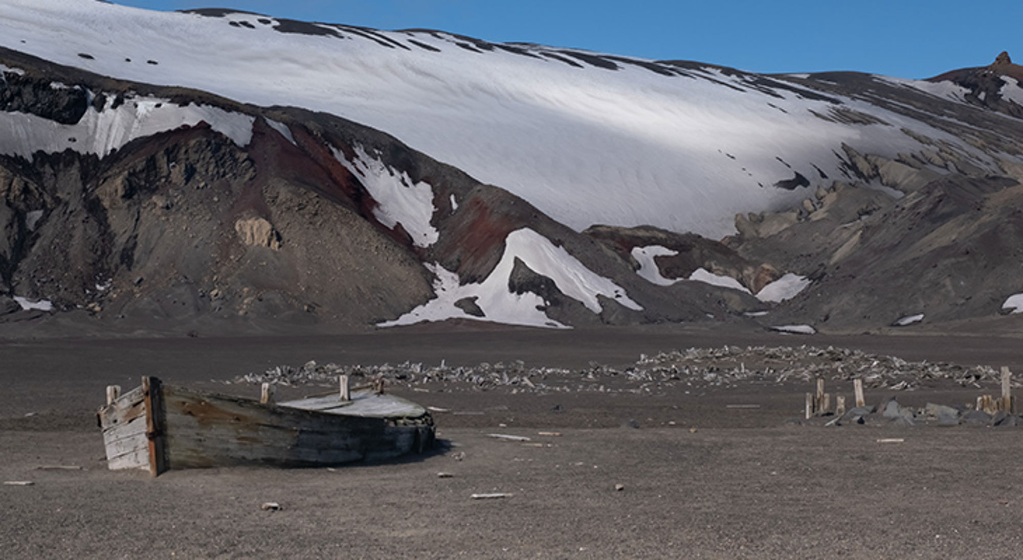 deception-island-krater Verlaten houten boot op zwart zand met besneeuwde bergen op de achtergrond.