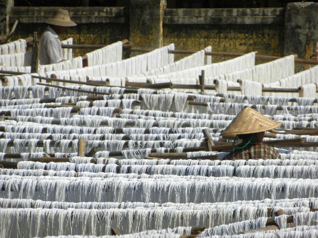 Witte textielstrengen drogen op houten rekken in Myanmar, met twee arbeiders tussen de rijen.