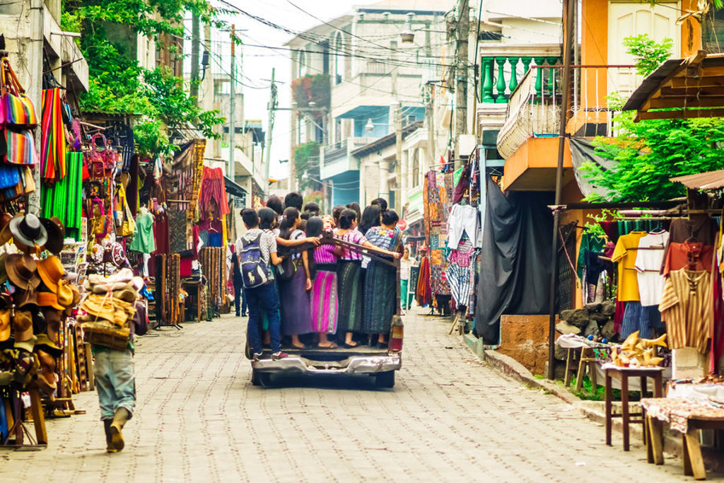Lokale marktstraat in Guatemala met kleurrijke textielkramen en mensen op de achterklep van een pick-up.