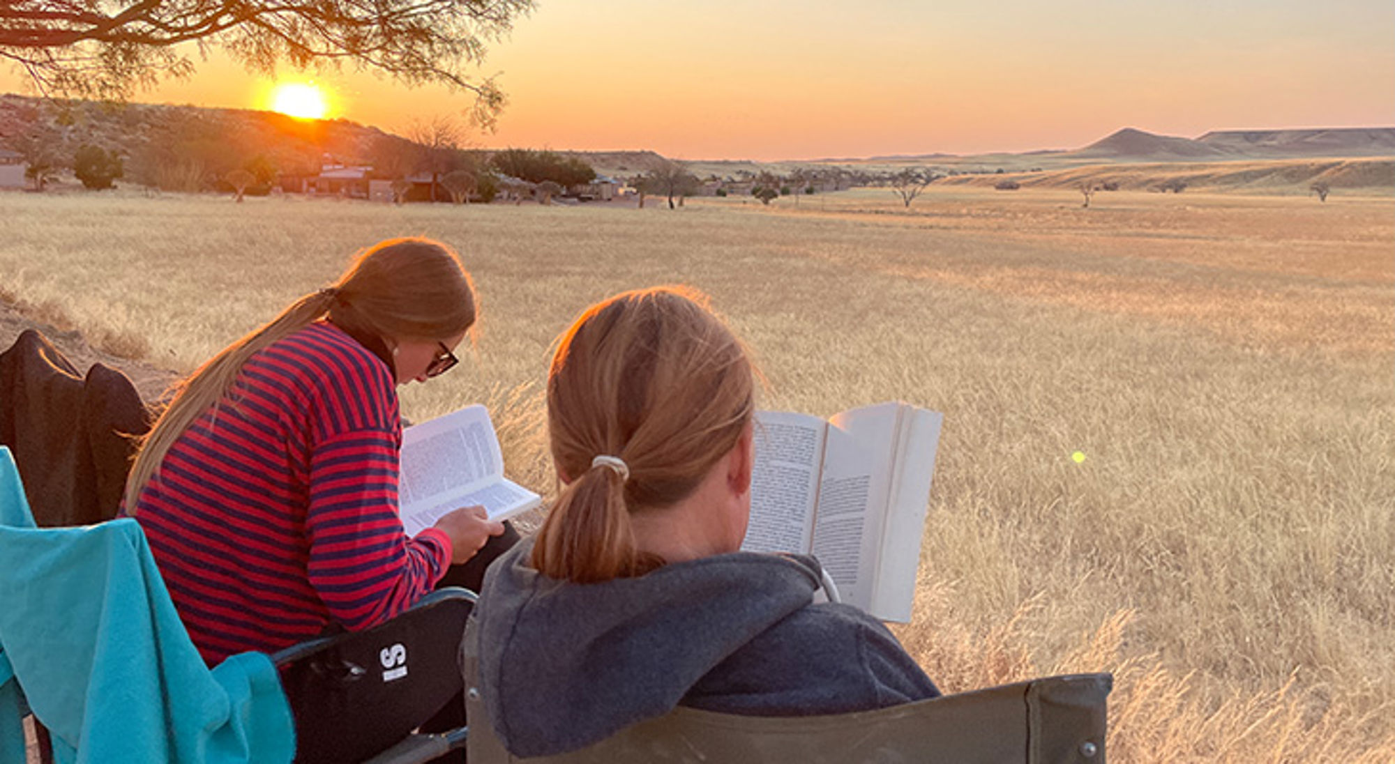 Familiereis Nambië, Botswana & Zimbabwe Twee reizigers lezen in kampeerstoelen tijdens zonsondergang op een savanneachtig grasland.