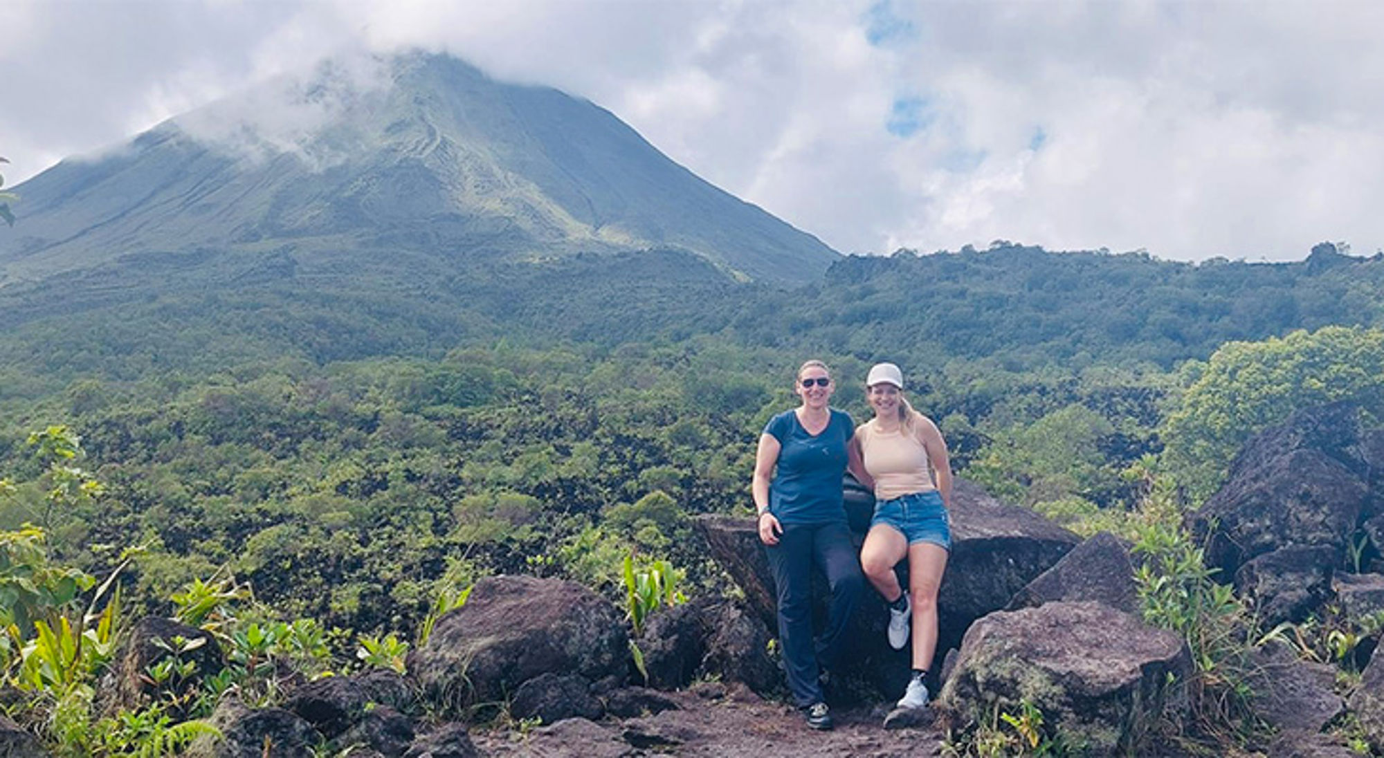 Sylvia en Lianne in Costa Rica Twee wandelaars poseren op rotsen met een vulkaan en groen regenwoud op de achtergrond.