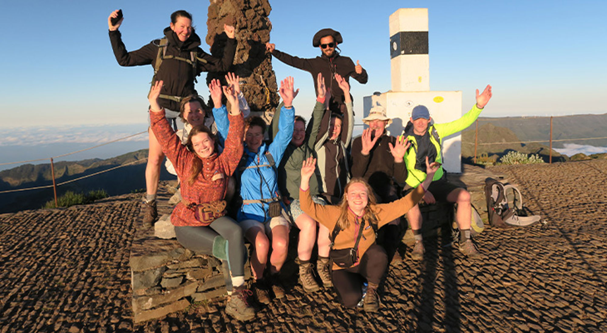 Groep wandelaars viert de top op een berg bij een geodetisch meetpunt, met uitzicht over het landschap.