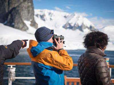 Reizigers spotten wildlife met verrekijker vanaf het dek, met besneeuwde bergen in Canada en Groenland op de achtergrond.