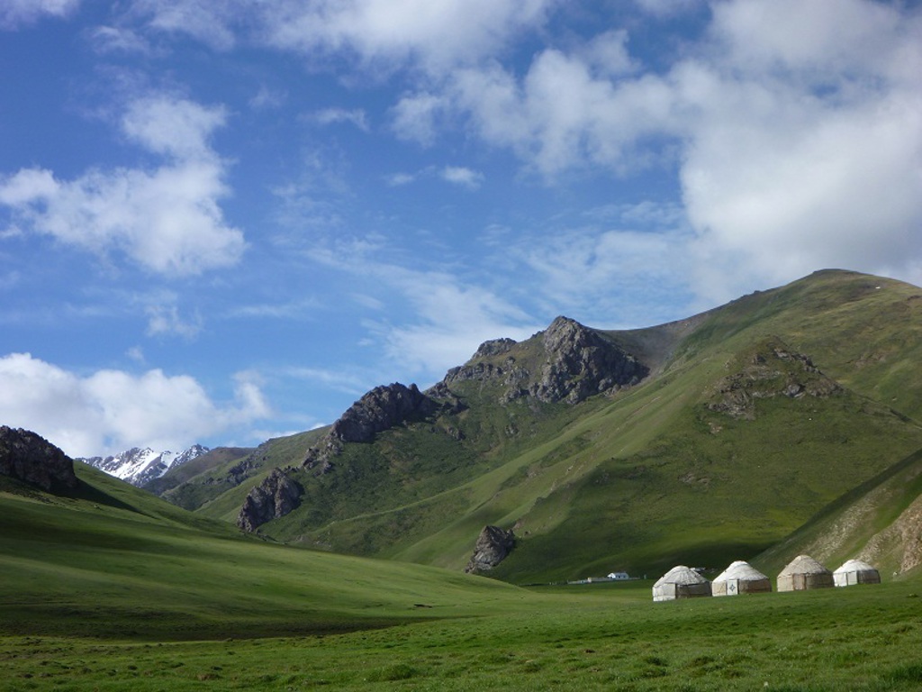 Groene bergvallei op de Zijderoute met yurts en besneeuwde bergtoppen onder een blauwe lucht.