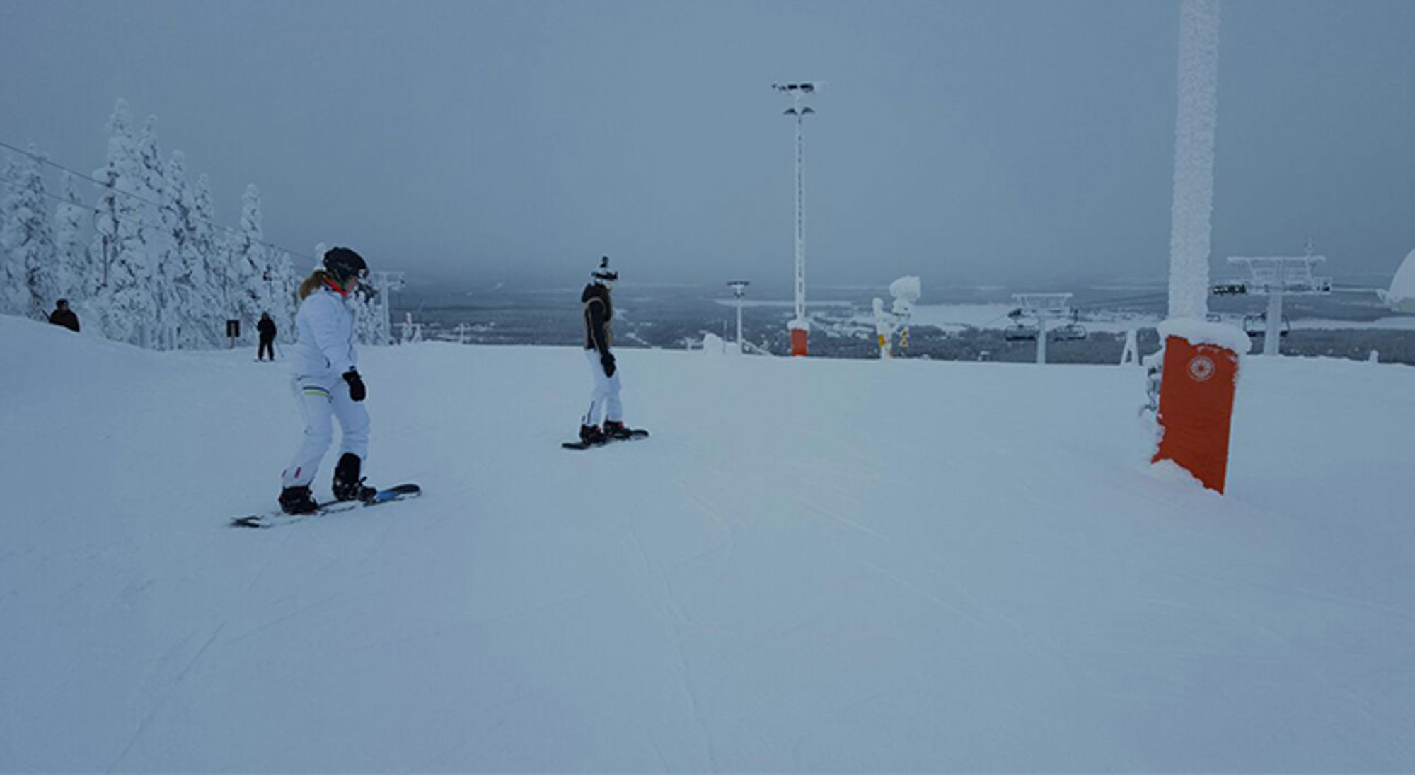 Snowboarders op een brede skipiste met besneeuwde bomen en uitzicht over het winterlandschap.