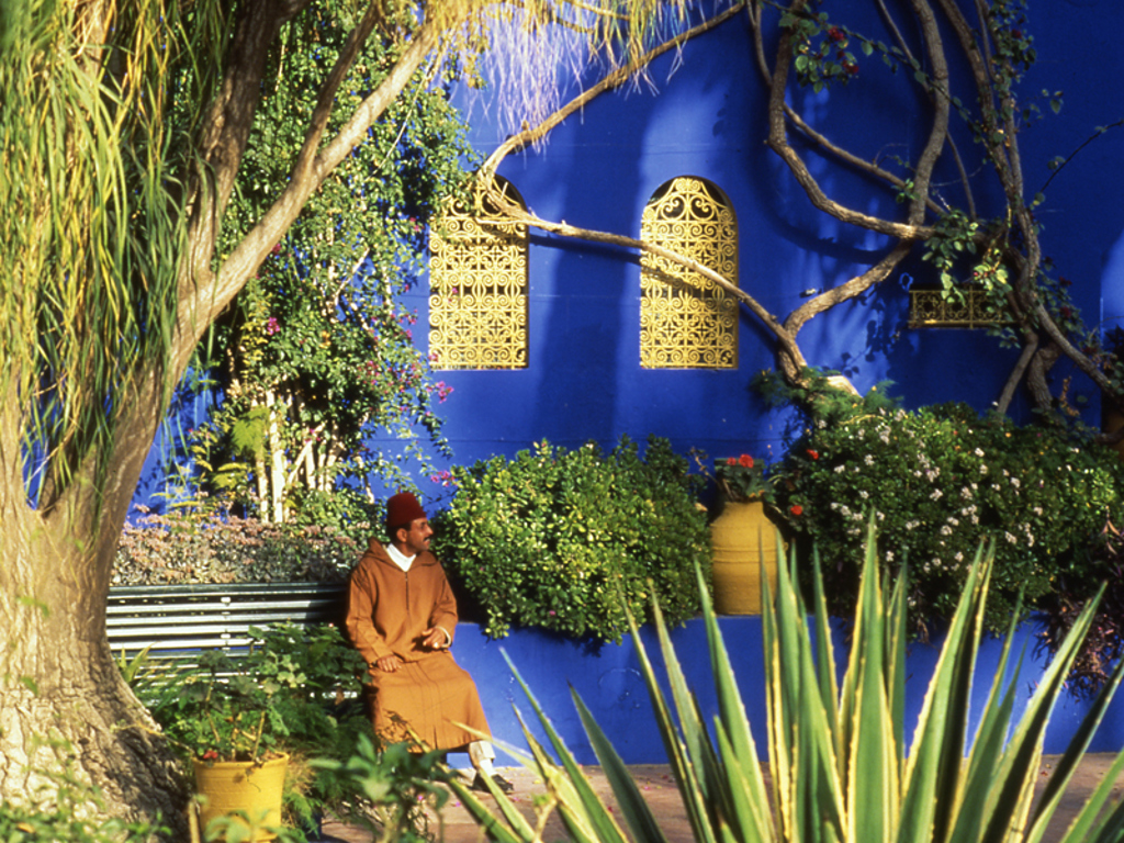 Man in traditionele kleding zit in de Majorelletuin in Marrakech, met blauwe muur en gele ramen op de achtergrond.