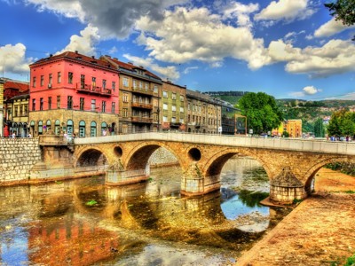 Stenen boogbrug in Sarajevo over de Miljacka, met kleurrijke gevels langs de kade.