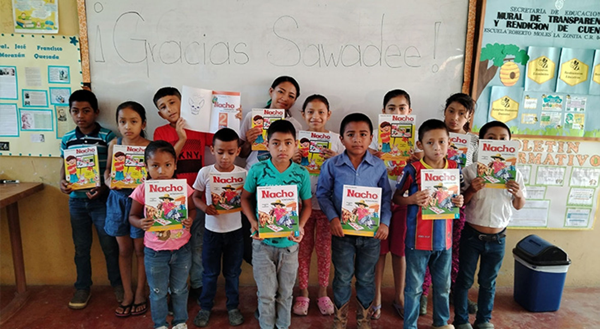 Schoolklas met leraar en leerlingen die lesboeken vasthouden voor een bord met de tekst “Gracias Sawadee!”.