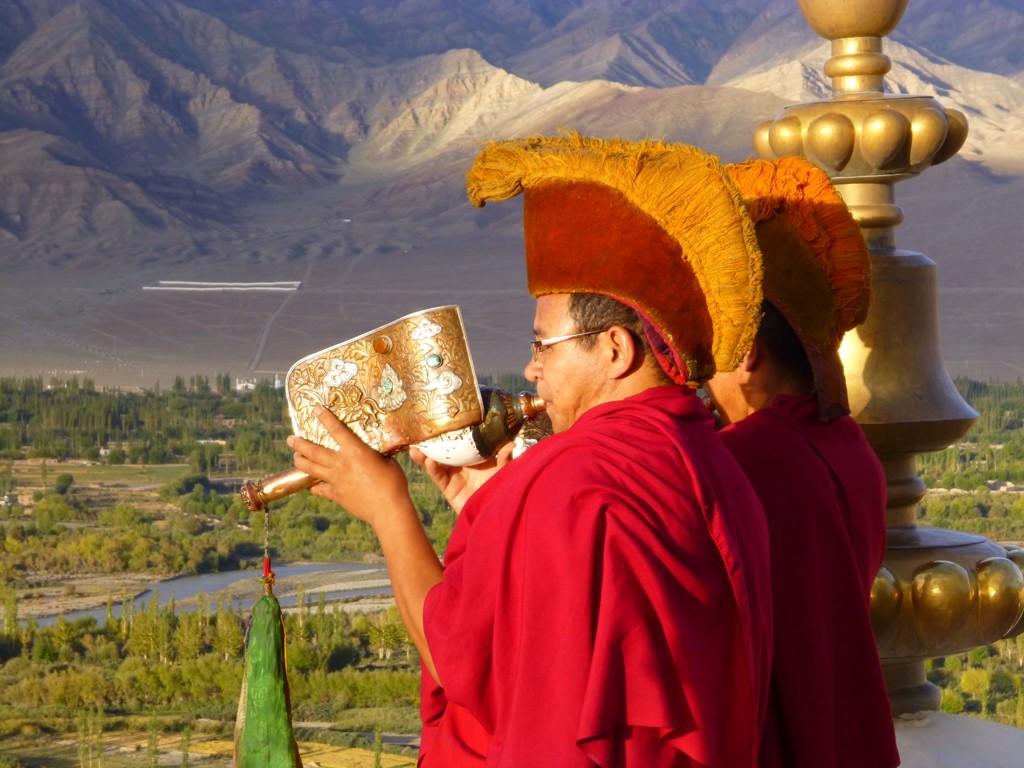 Boeddhistische monniken blazen op een rituele hoorn bij een klooster in Ladakh, met bergen op de achtergrond.