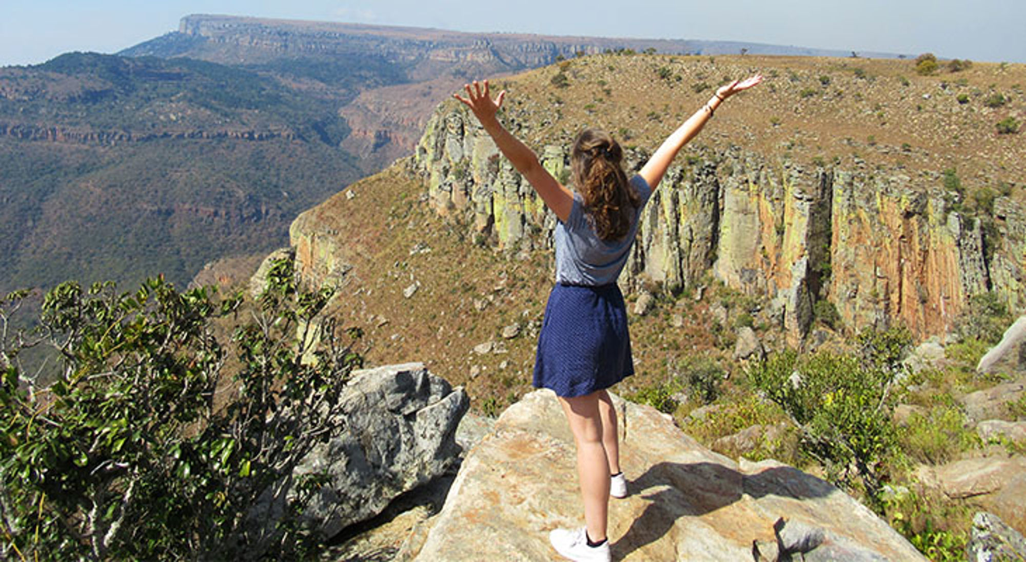 Vrouw met opgeheven armen op rots met uitzicht over een bergkloof en plateau.