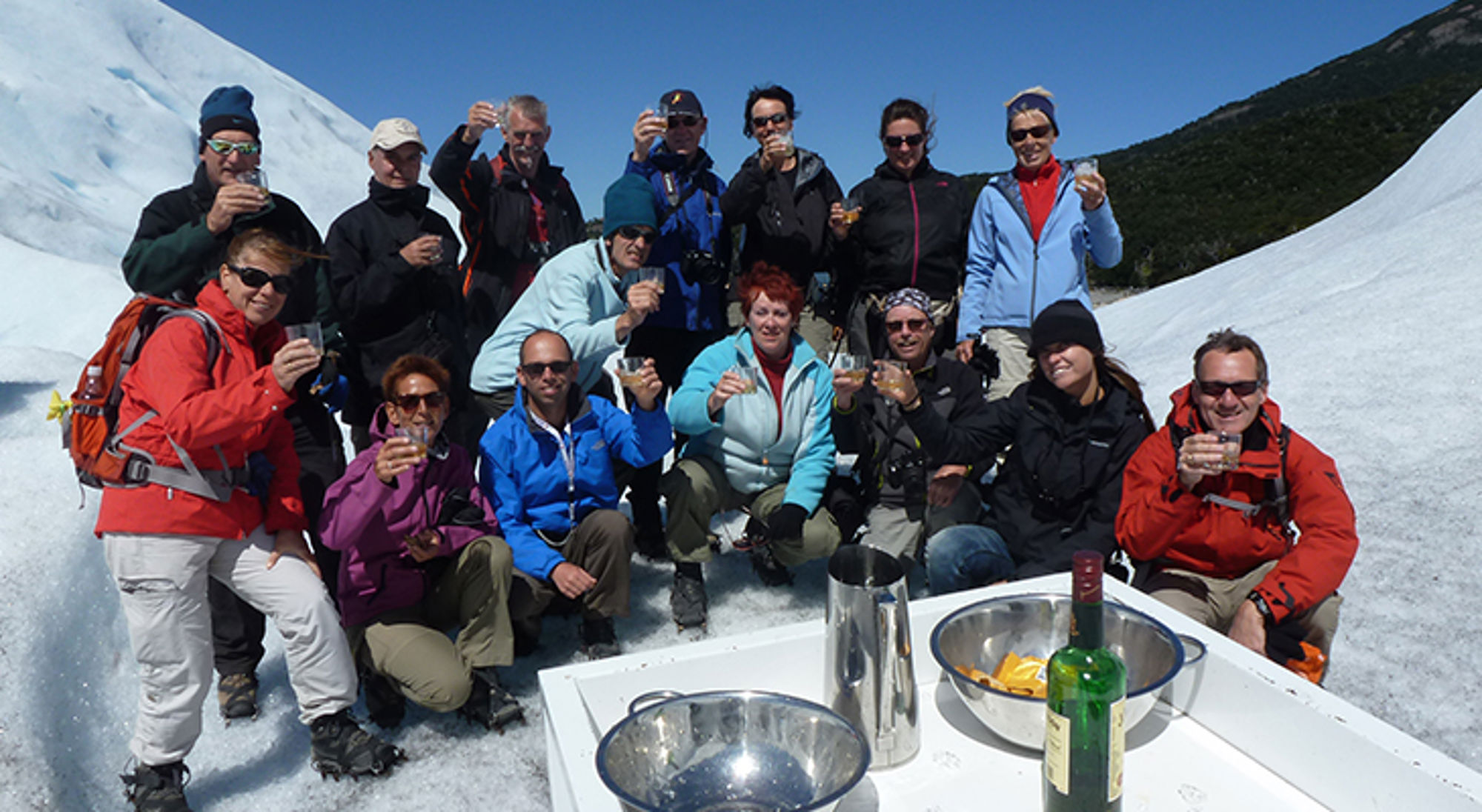 Groep wandelaars proost met glazen op een gletsjer, met tafel en drank op de voorgrond.