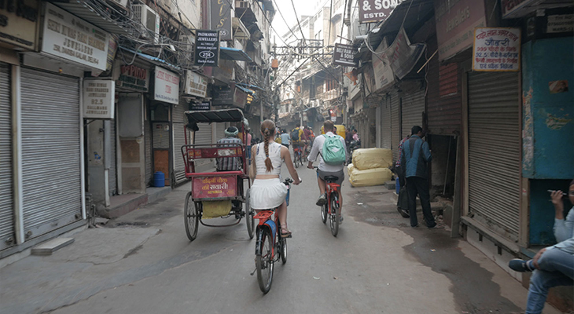 Fietsers in een smalle straat met riksja’s en winkels in Delhi, India.