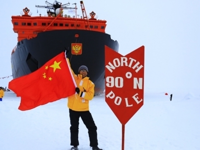 Reiziger met vlag bij het bord ‘North Pole 90°N’ op het ijs, met ijsbreker op de achtergrond.