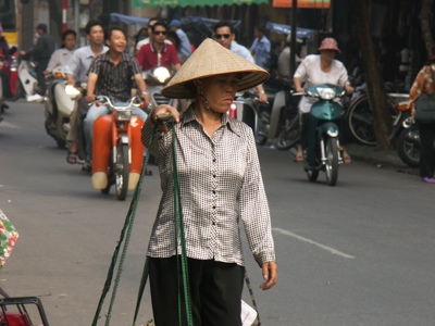 Straatbeeld in Hanoi met vrouw met rieten hoed die manden draagt, tussen scooters en druk verkeer.
