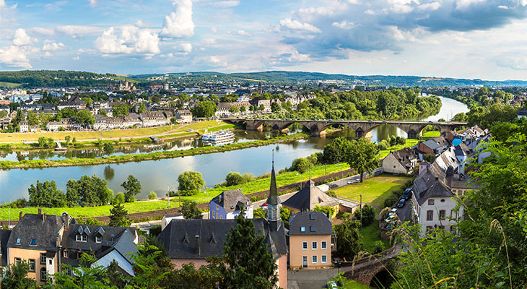 Bernkastel-Kues Panoramisch uitzicht op een rivier met brug, groen landschap en stad met kerk in de verte.