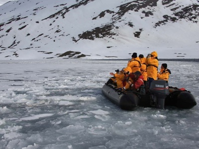Zodiacboot met expeditieleden in oranje jassen vaart door drijfijs langs besneeuwde kust tijdens Noordpoolrondreis.