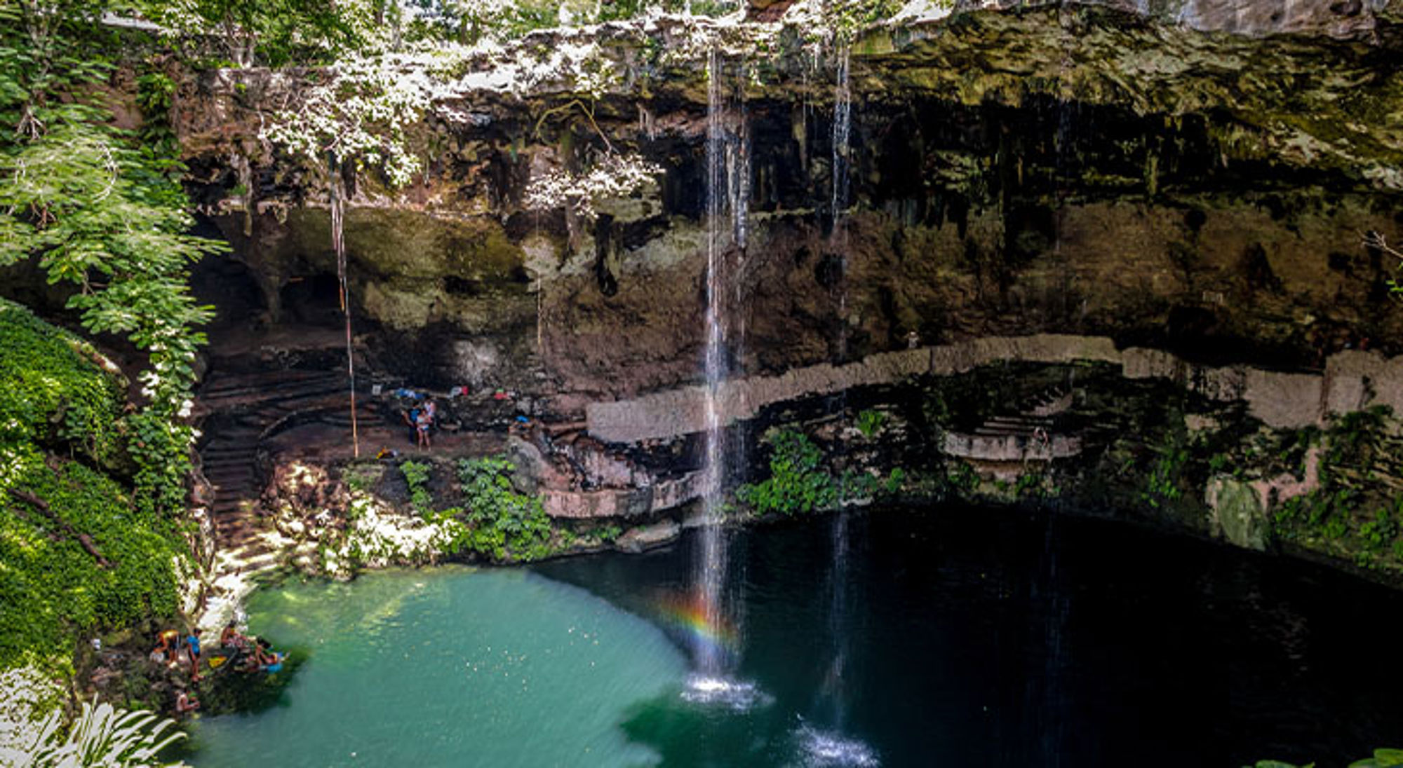 Cenote Zaci Waterval in een grot die uitkomt in een turquoise cenote, omringd door groen.