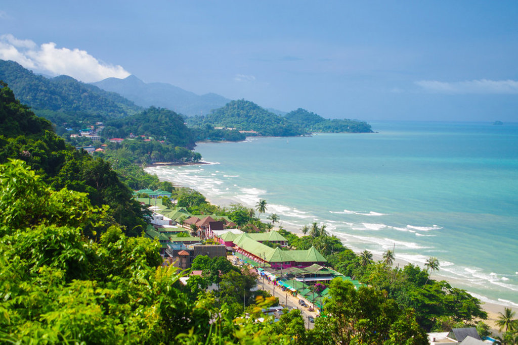 Uitzicht over een tropische kust met strand, palmbomen en groene bergen tijdens een familiereis Thailand, Laos en Cambodja.