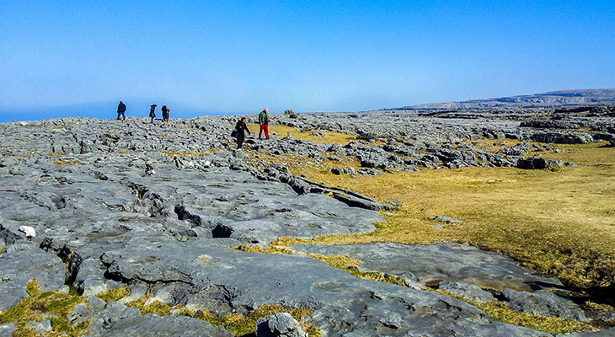 The Burren, Ierland Wandelaars op een rotsachtig kustlandschap onder een strakblauwe lucht.