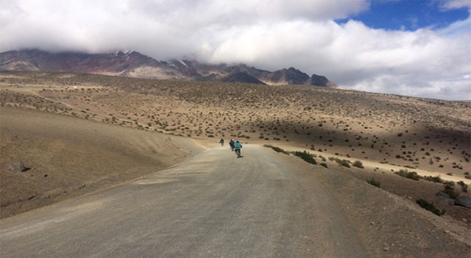 Volcán Chimborazo Motorrijders op een lege bergweg door een droog landschap met donkere bergtoppen en wolkenlucht.