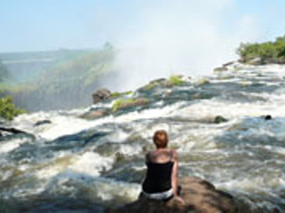 Reiziger bij de rand van Victoria Falls, met opspattend water en regenboog in de nevel.