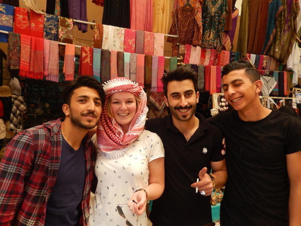 22-35ers reis Jordanië: reiziger poseert met locals op een markt met kleurrijke sjaals op de achtergrond.