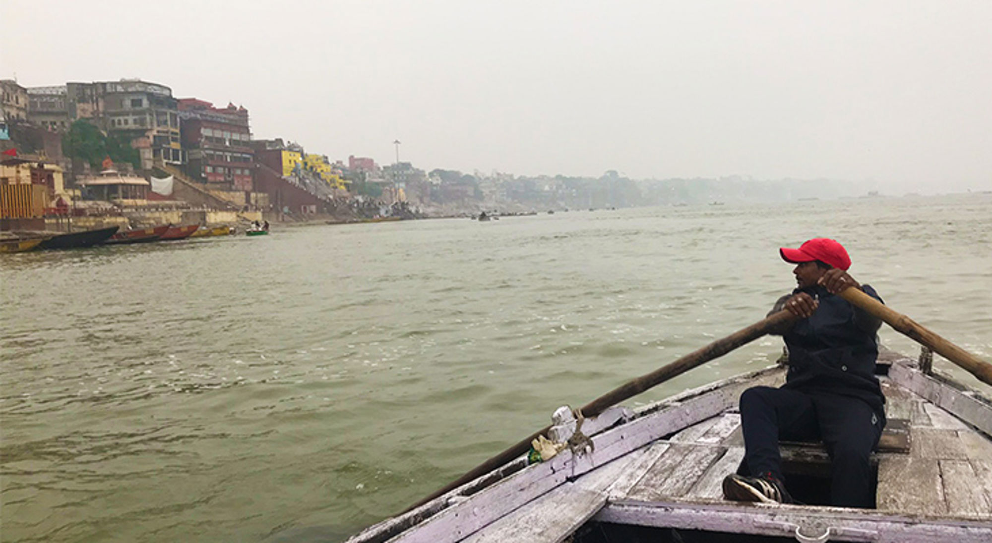 Man roeit in een houten boot over de Ganges langs de ghats van Varanasi.