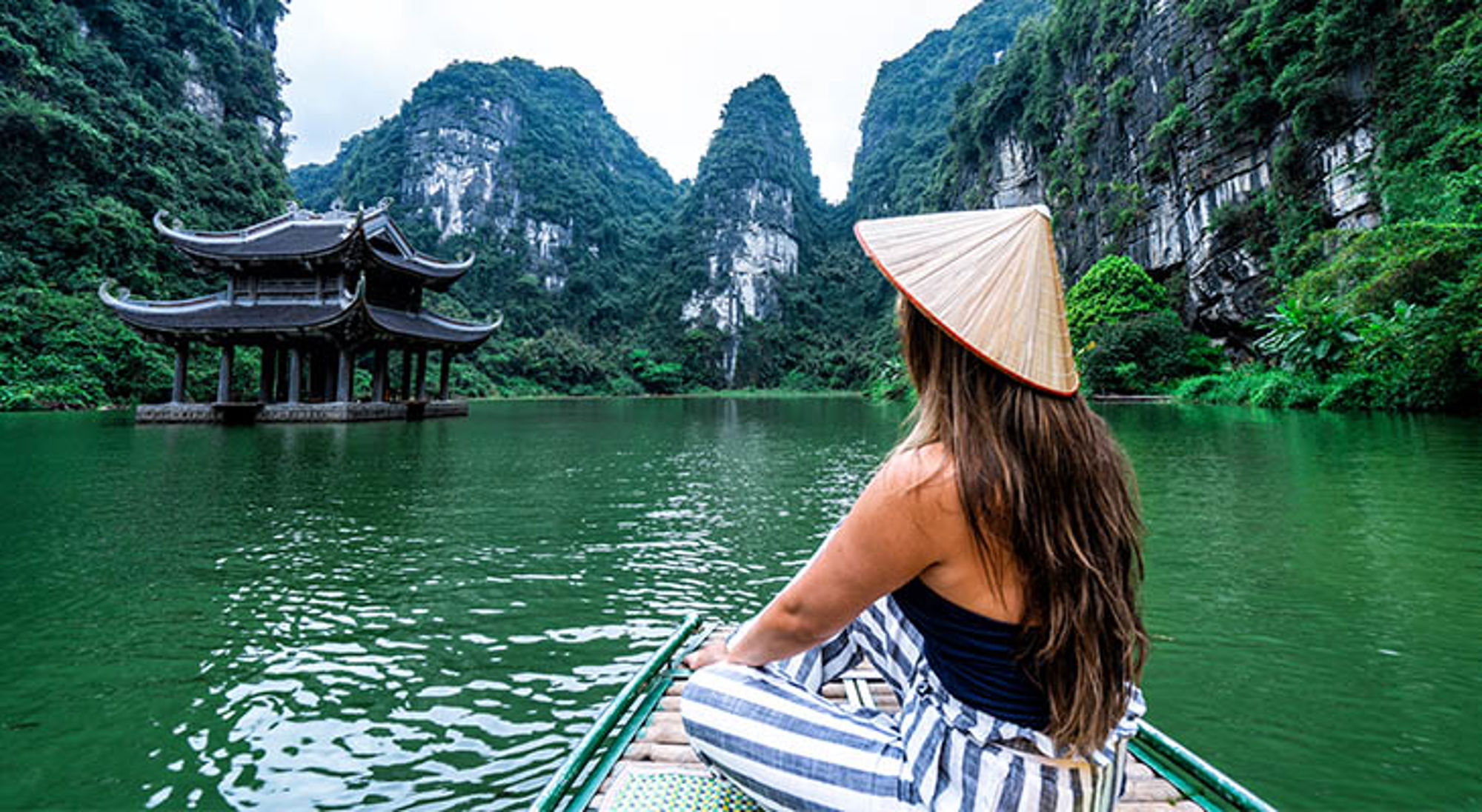 Vrouw met Vietnamese hoed op een bootje bij Trang An, Ninh Binh, met pagode en kalksteenbergen.